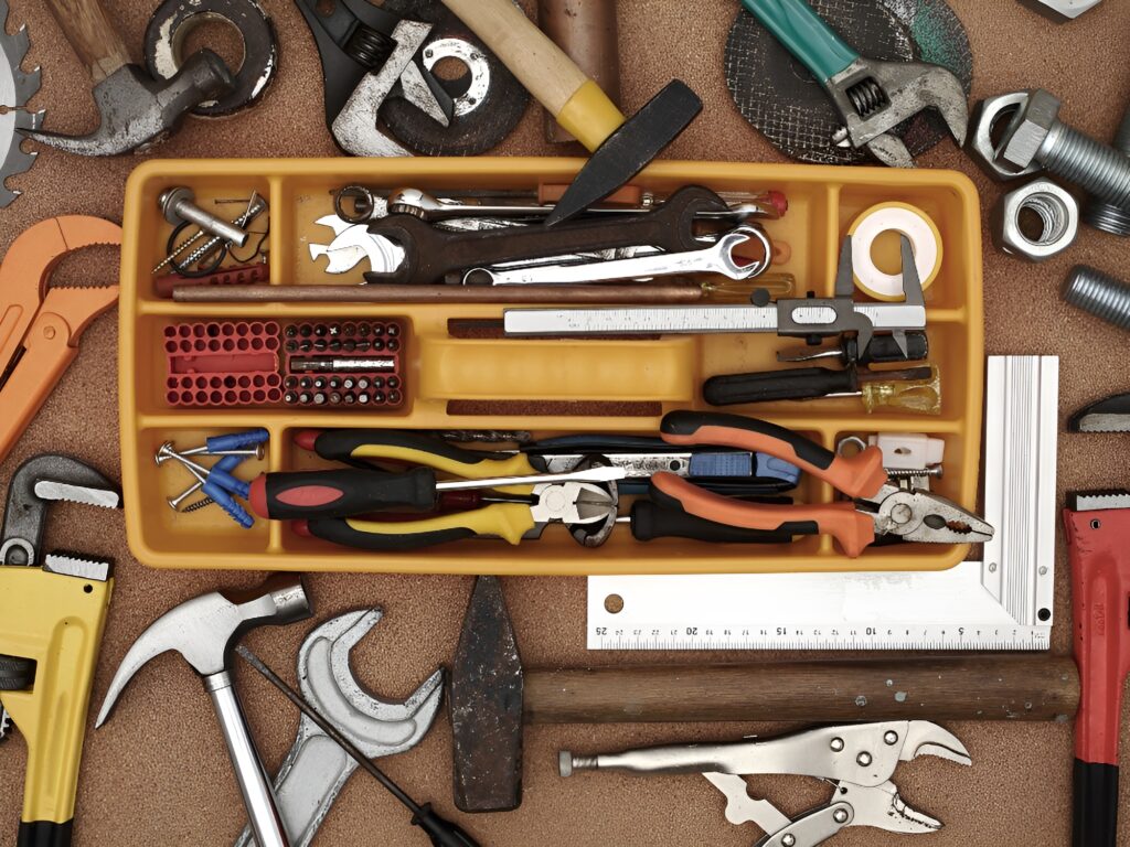 An extensive array of DIY tools for homeowners is meticulously laid out on a sturdy workbench. Key items in this essential home improvement collection include a shiny claw hammer, versatile adjustable wrenches, a precision utility knife with a conspicuous yellow handle, and an assortment of screwdrivers suitable for various tasks. Organized within a vibrant yellow tool tray are neatly aligned screwdriver bits in a compact red case, a handy retractable measuring tape, and multiple sizes of wrenches. Additional items interspersed on the work surface are assorted screws, hefty nuts, solid bolts, a circular saw blade, a reliable spirit level, and a characterful, aged hatchet, all signifying the comprehensive nature of the basic DIY toolkit necessary for adept homeowners.